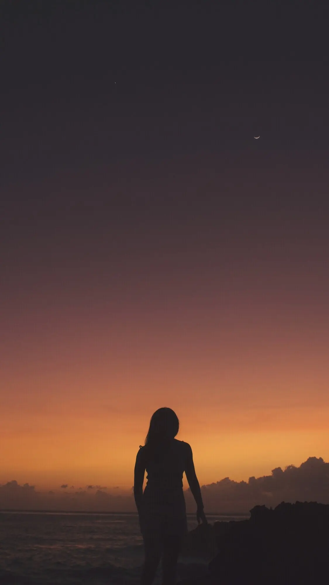 Hochformataufnahme einer Frau als Silhouette, die bei Sonnenuntergang auf einer Klippe nahe dem Meer steht.